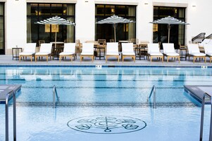 Une piscine entourée de chaises longues et de tables, le tout adossé à un bâtiment doté de grandes fenêtres.