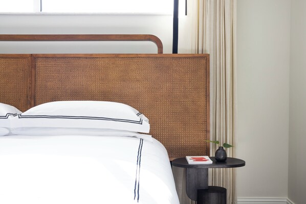 A neatly made bed with white linens and dark trim, a wooden headboard, and a small round table with a book and a vase. - Kimpton Cottonwood by IHG (Omaha)