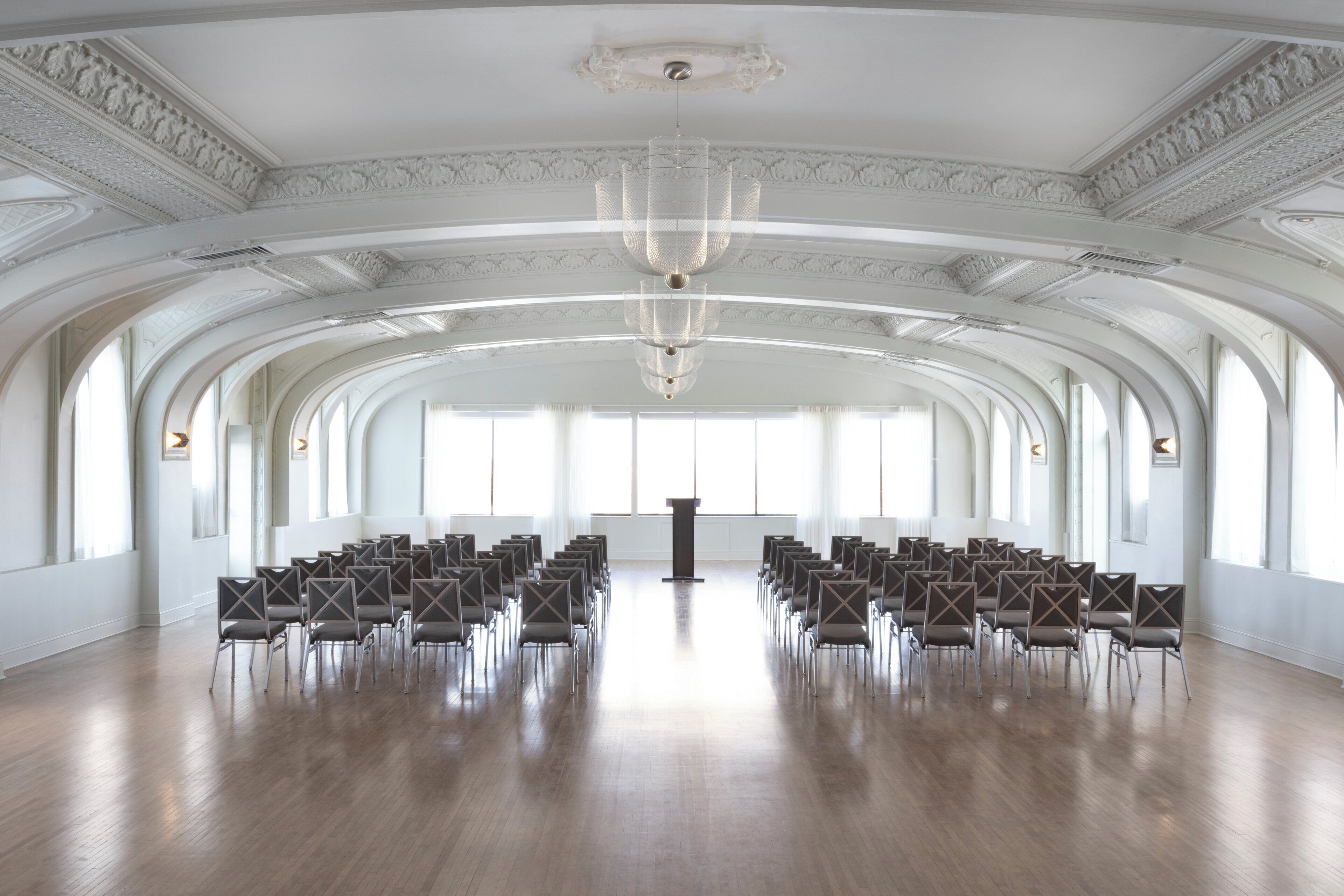 Een ruime hal met een hoge plafond, boogvensters en rijen stoelen, klaar voor een evenement.