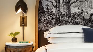 A neatly made bed with white linens and a blue pillow, a wall-mounted lamp, a small round side table with a plant and books, and a large tapestry depicting a tree and landscape.