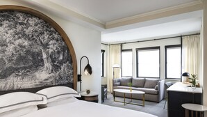 A hotel room with a large bed, a sofa, a small table, and a desk. There are windows with curtains and a wall-mounted lamp. - Kimpton Cottonwood by IHG (Omaha)