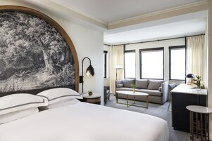 A hotel room with a large bed, a sofa, a small table, and a desk. There are windows with curtains and a wall-mounted lamp.
