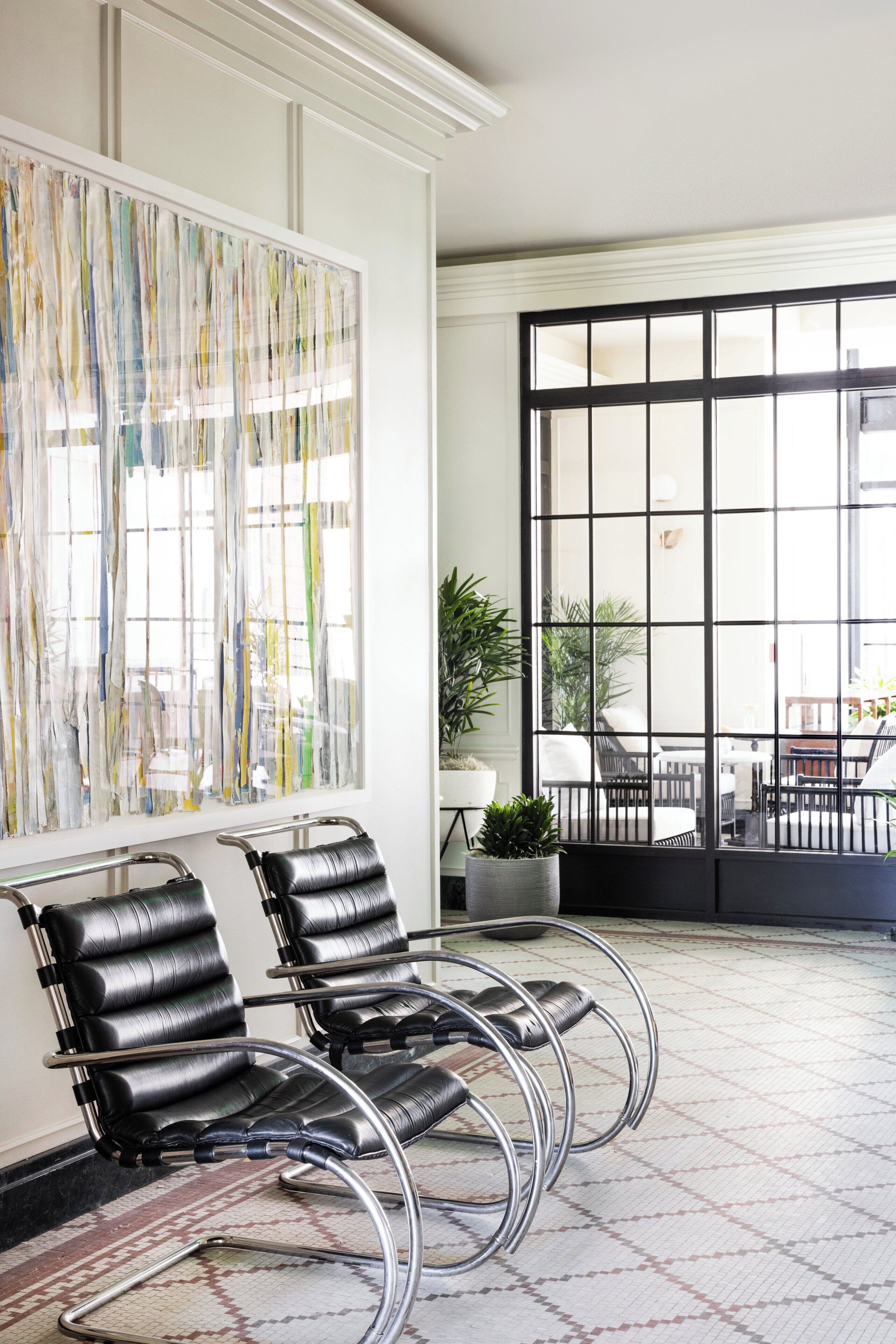 A modern interior with a seating area featuring black leather chairs with metal frames, a large abstract painting on the wall, and a view through a large window to an outdoor seating area.