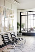 Un intérieur moderne avec un espace salon comprenant des fauteuils en cuir noir à structure métallique, une grande peinture abstraite au mur et une vue, par le biais d’une grande fenêtre, sur un espace salon extérieur.