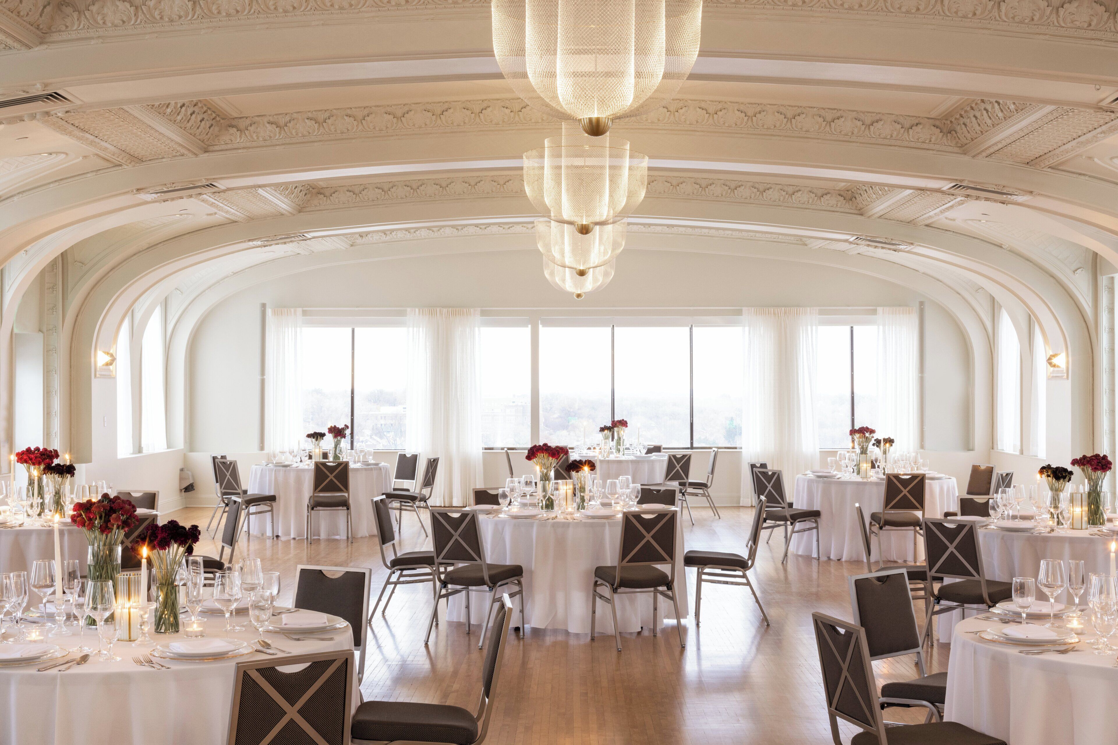 Een ruime eetzaal met hoge plafonds, sierlijke ornamenten en grote ramen. Tafels zijn gedekt met witte tafelkleden, stoelen en bloemstukken.