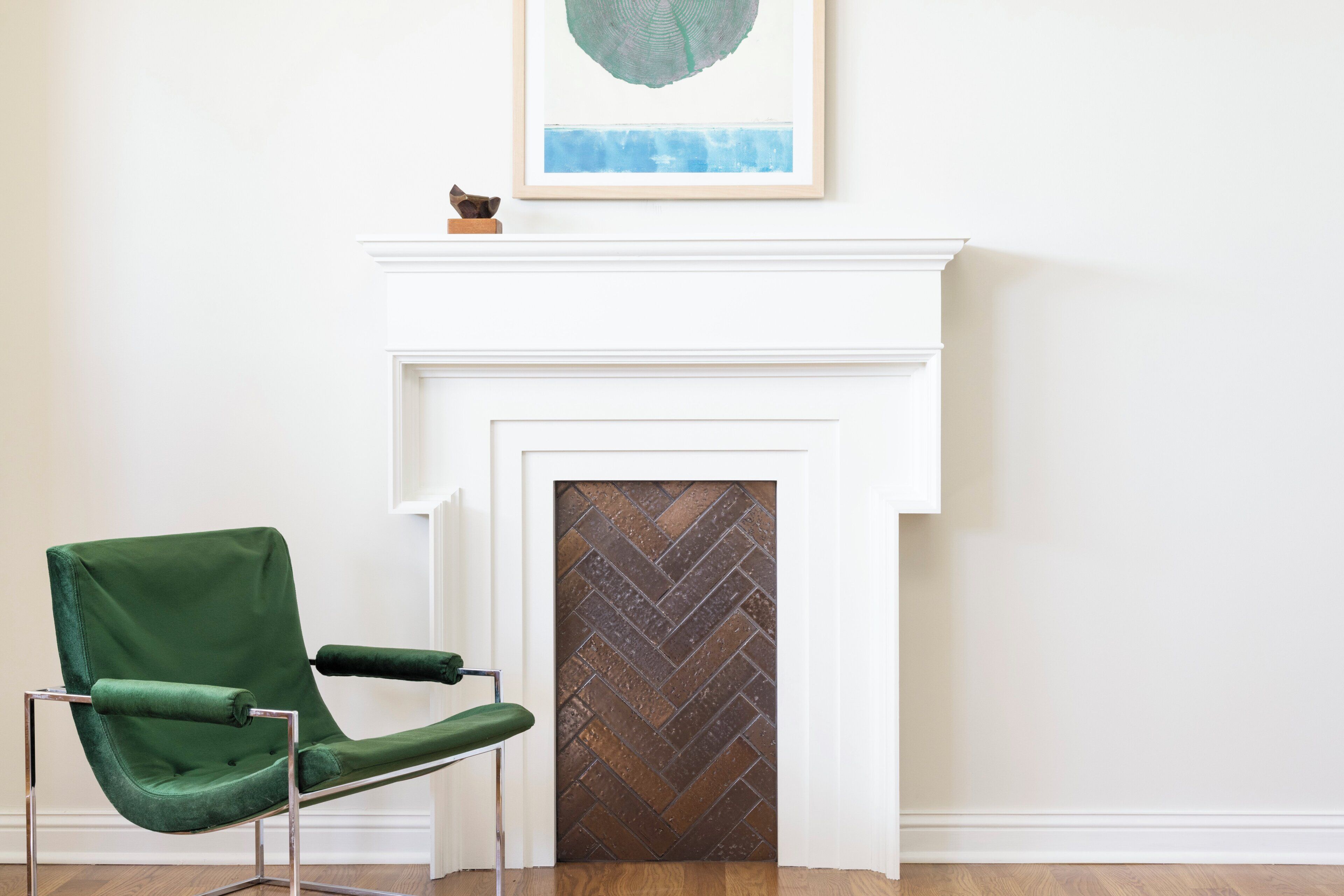 A room with a green chair, a white fireplace, and a framed artwork above it.
