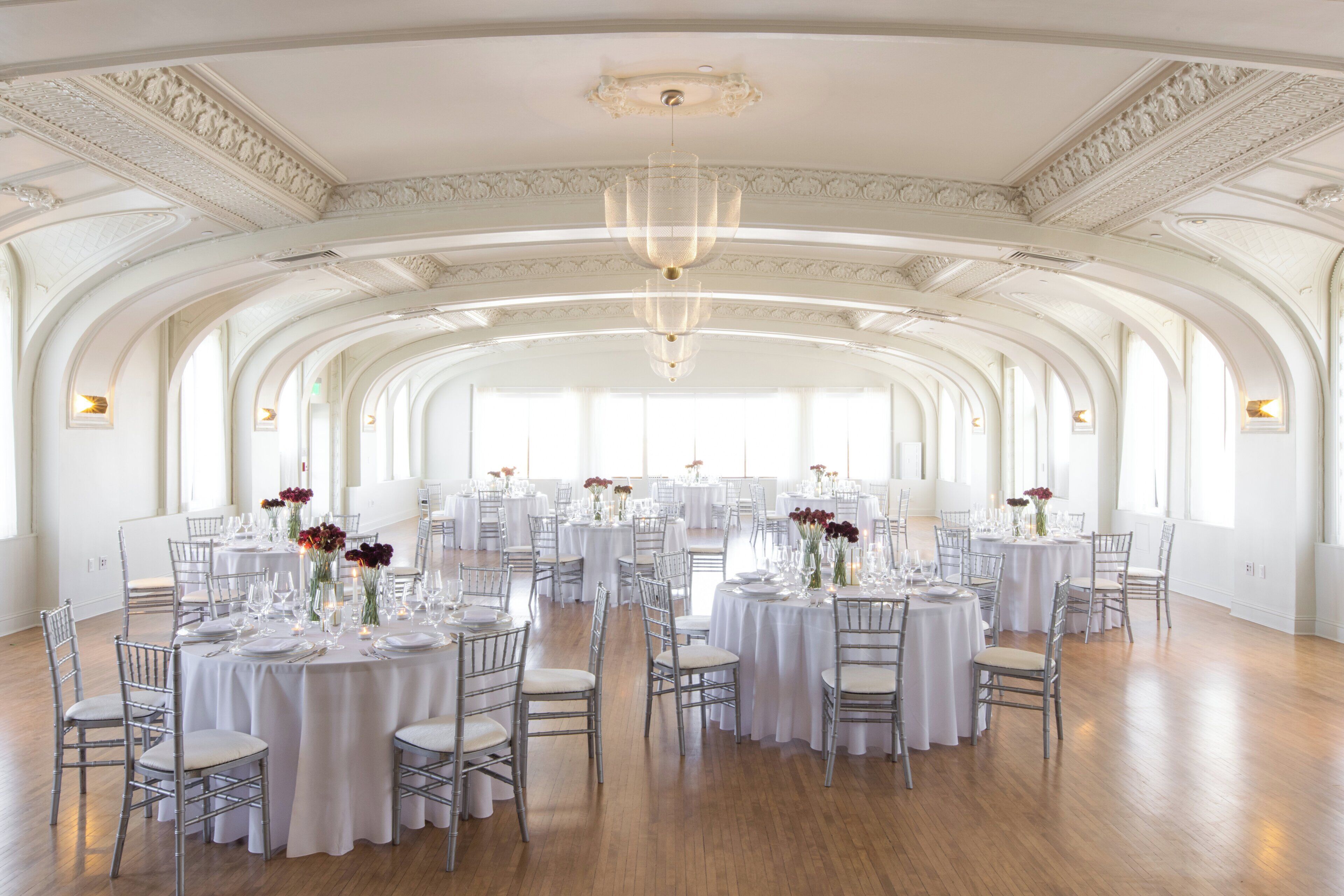 A spacious hall with high ceilings and arched windows, furnished with round tables and chairs, and decorated with floral centerpieces.