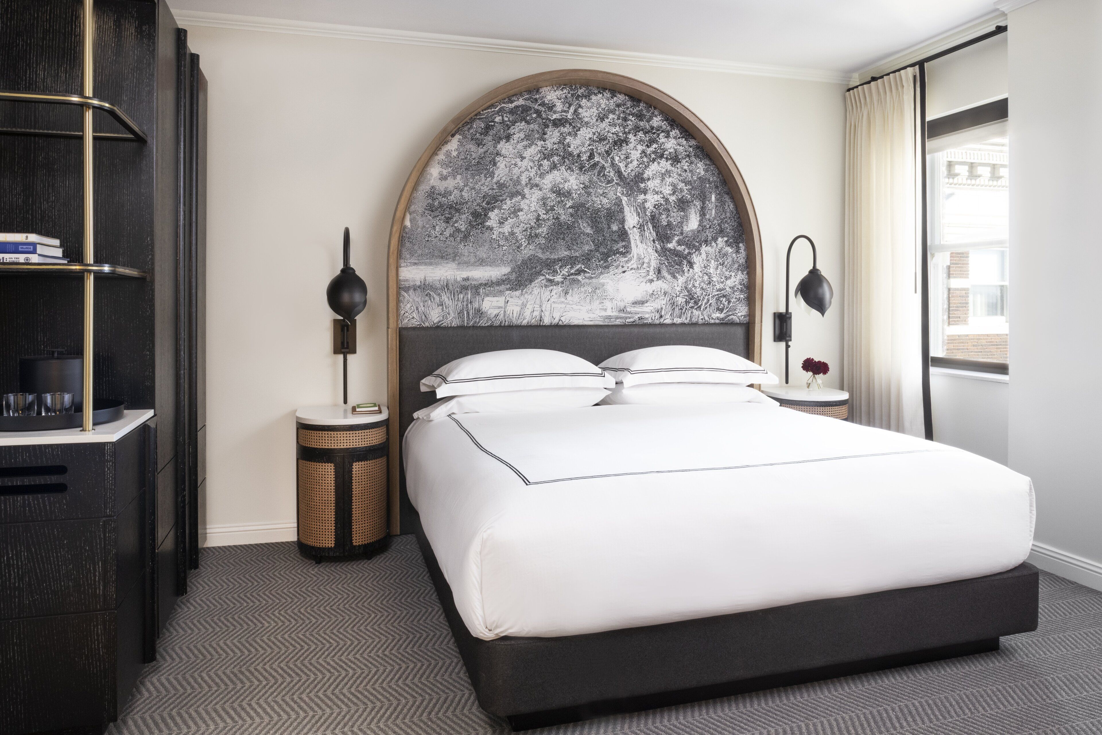 A hotel room with a large bed, a round headboard with a nature scene, bedside tables with lamps, a bookshelf, and a window with curtains.