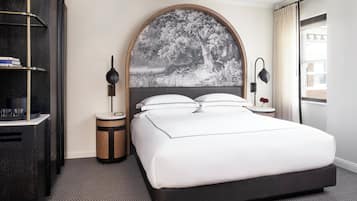 A hotel room with a large bed, a round headboard with a nature scene, bedside tables with lamps, a bookshelf, and a window with curtains.