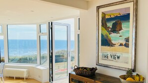 A spacious room with a large sliding glass door leading to a balcony with a view of the sea. There is a framed poster on the wall and a wooden cabinet with drawers. - Luxury, Spacious, Beachfront (direct access) House with Breath-taking Sea Views (Downderry)