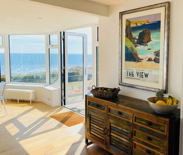 Ruime kamer met een grote schuifpui die uitkomt op een balkon met uitzicht op zee. Er hangt een ingelijste poster aan de muur en staat een houten kast met lades.