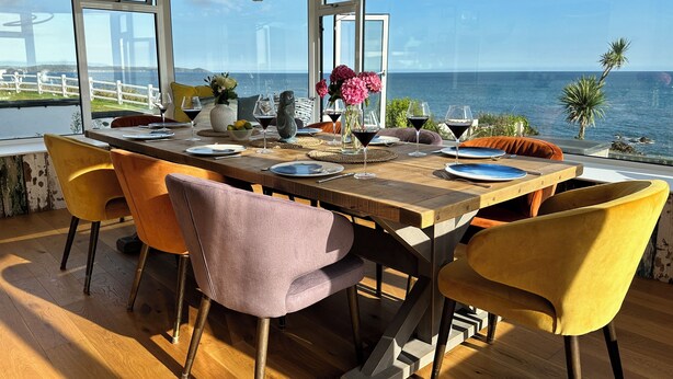 A dining area with a view of the sea, wooden table, and colorful chairs. - Luxury, Spacious, Beachfront (direct access) House with Breath-taking Sea Views (Downderry)