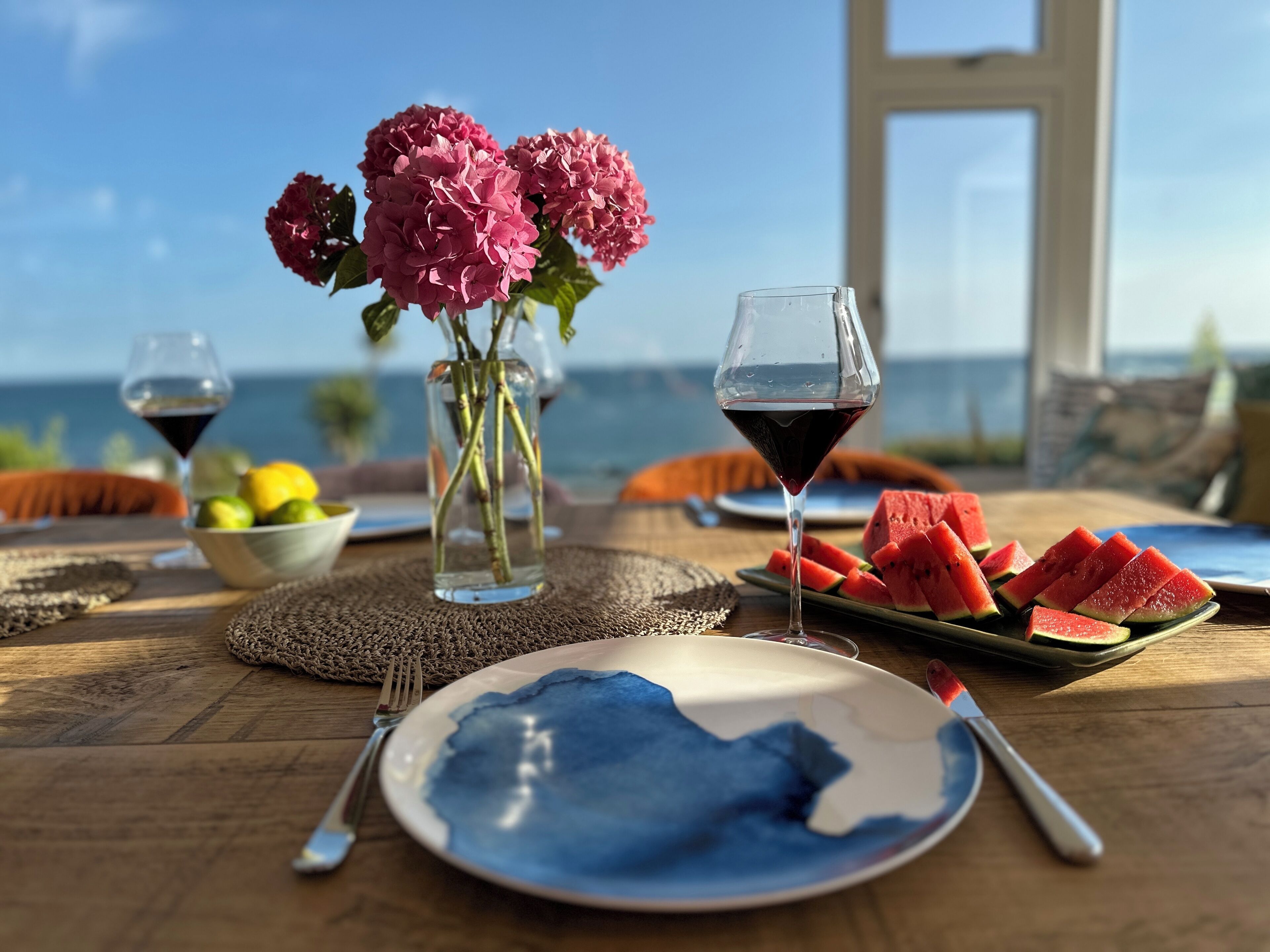 Una mesa con una porción de sandía, una copa de vino tinto y un jarrón con flores.