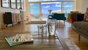 A spacious living room with a large window offering a view of the sea, a wooden coffee table with magazines and glasses, and a cozy seating area with a sofa and armchairs. - Luxury, Spacious, Beachfront (direct access) House with Breath-taking Sea Views (Downderry)