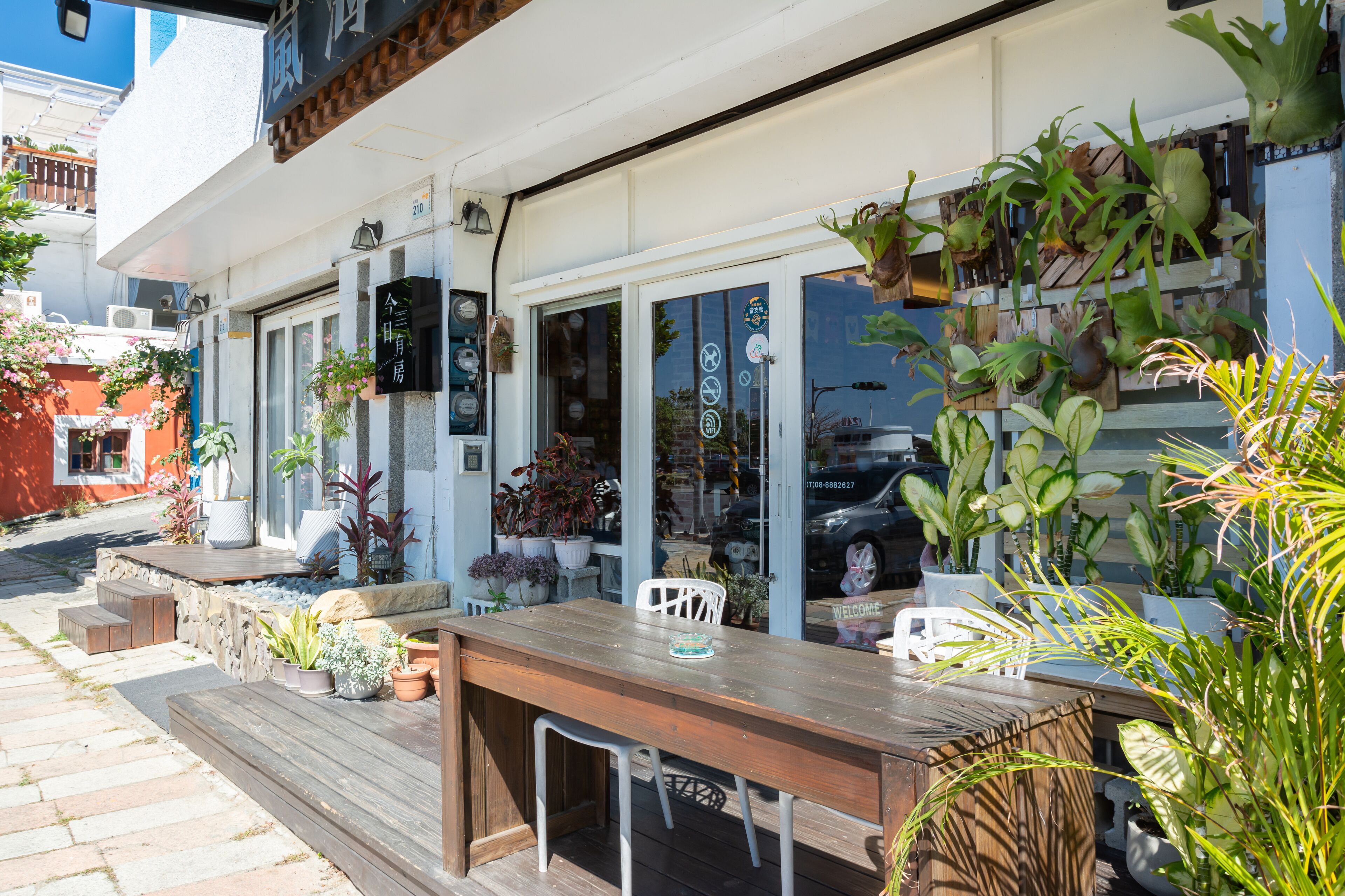 An outdoor patio with a wooden table and chairs, surrounded by potted plants and a variety of greenery.