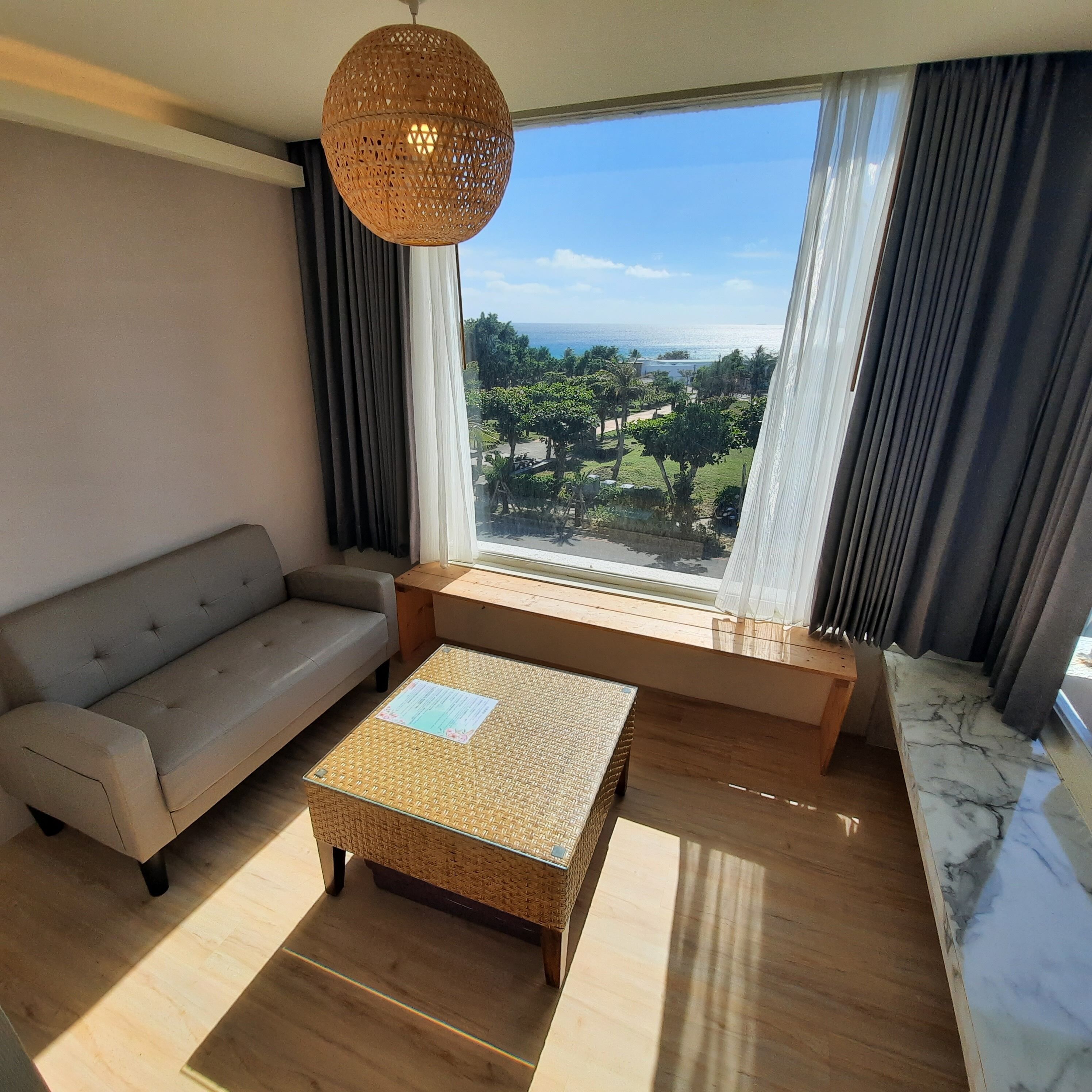 Un salon moderne avec un canapé, une table basse en osier et une vue sur un paysage côtier, visible à travers de grandes fenêtres.