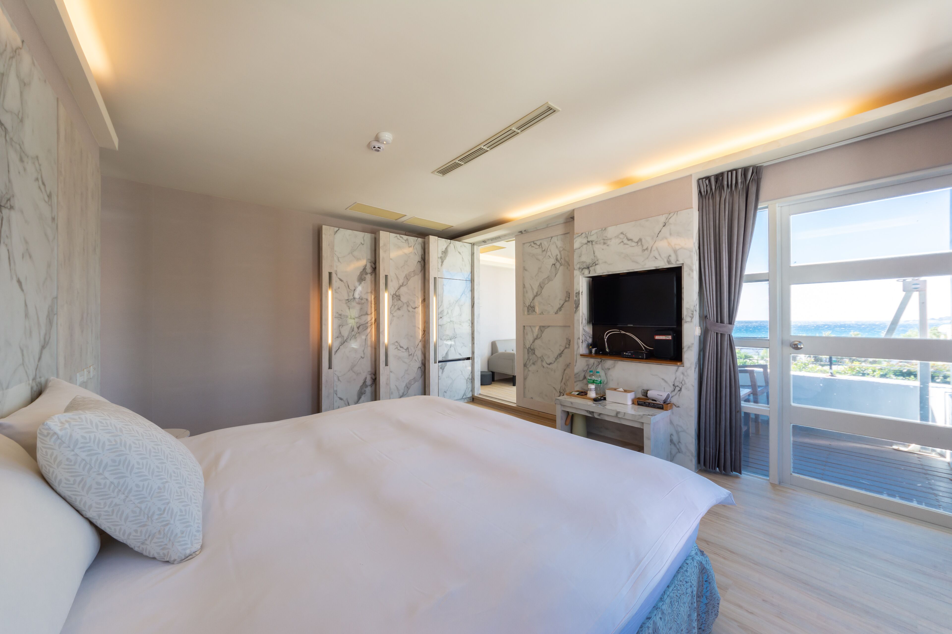 A modern hotel room with a large bed, marble walls, a TV, and a balcony with a view.