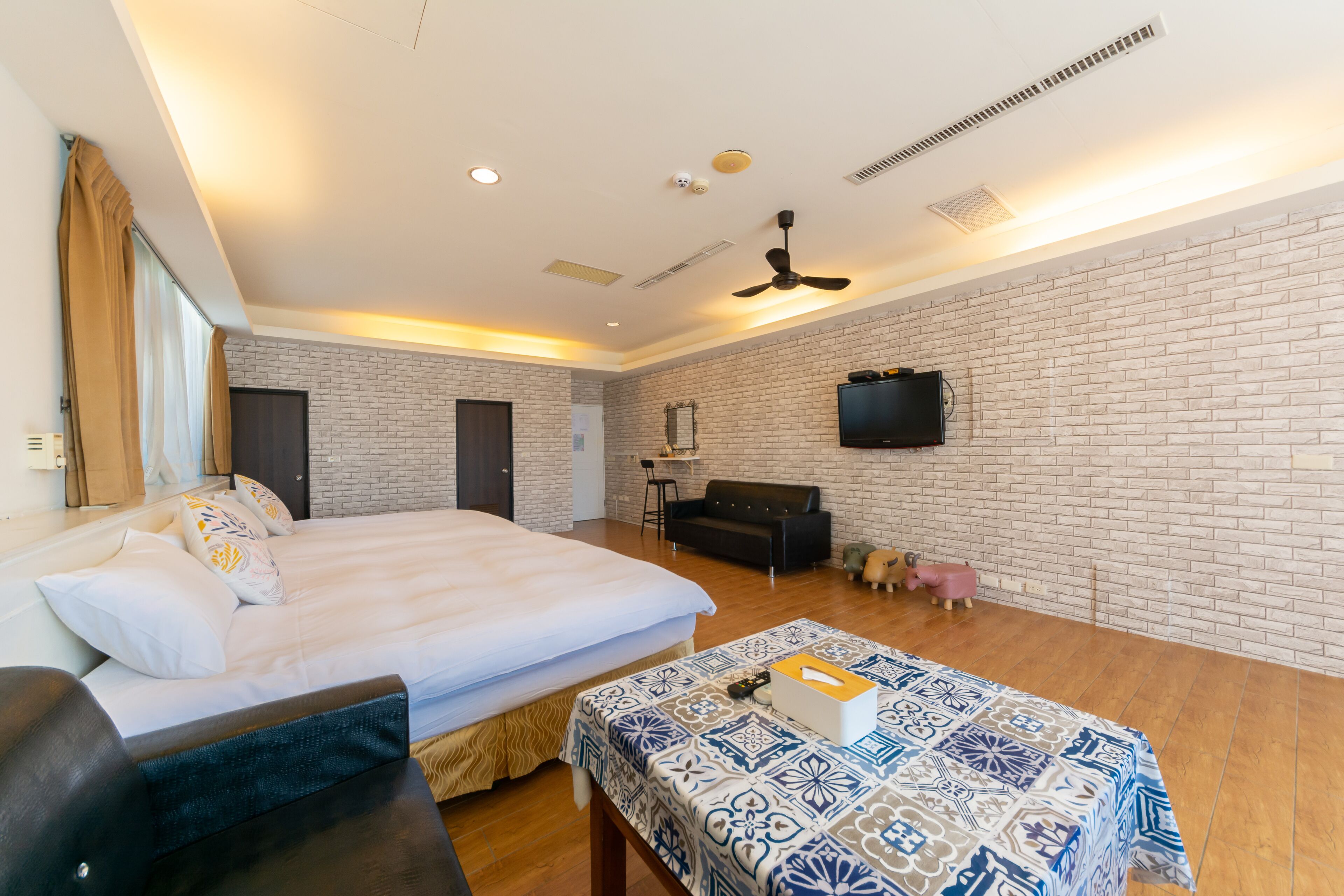 A modern hotel room with a large bed, a sofa, a dining table, and a TV mounted on the brick wall.
