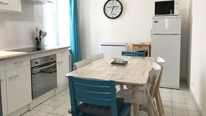 A compact kitchen with white cabinets, a microwave, and a refrigerator. There is a dining table with chairs, a window with curtains, and a wall clock. - Apartment historic center Neufchâteau (Neufchâteau)