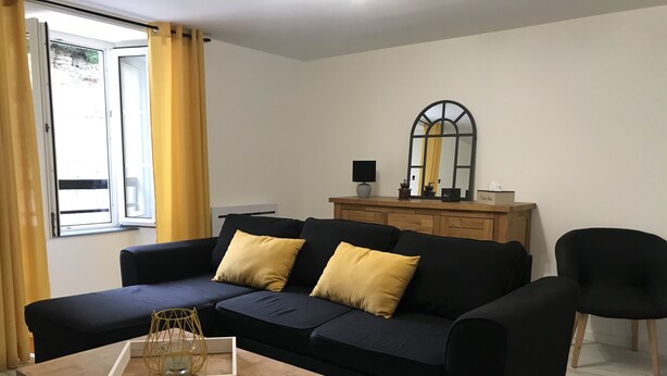A modern living room with a dark blue sofa, two mustard yellow cushions, a wooden coffee table with a decorative glass container, a black armchair, and a wooden cabinet with a mirror above it. - Apartment historic center Neufchâteau (Neufchâteau)