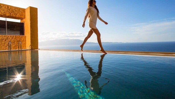 Eine Person geht über einen Pool mit Glasboden. Unter dem Pool ist das klare Meer zu sehen, im Hintergrund ein Gebäude mit einer Steinfassade.