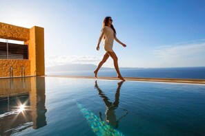A person walking on a glass-bottomed infinity pool with a clear view of the ocean and a building with a stone facade. - Eleni's Serenity - Discovering Sapphire Harmonies (Ravdoucha)