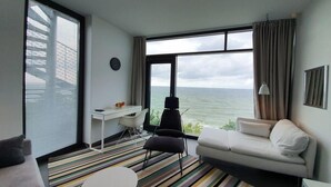 A modern hotel room with a large window offering a sea view, a white sofa, a black chair, a small desk, and a striped rug. - Apartamenty Boulevard - 365PAM (Ustronie Morskie)