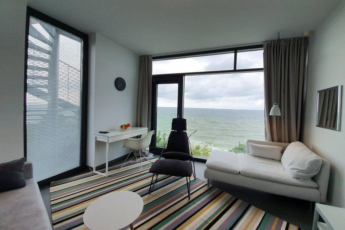 Habitación de hotel moderna con un amplio ventanal con vistas al mar, un sofá blanco, una silla negra, un escritorio pequeño y una alfombra a rayas.