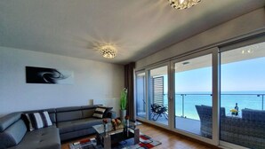 Ein modernes Wohnzimmer mit einer Couch, einem Couchtisch und Blick auf das Meer durch große Schiebegläser.