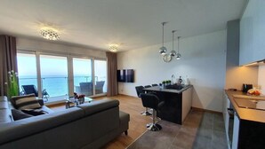 Ein modernes Wohnzimmer mit Sofa, Essbereich und Küche. Ein großes Fenster bietet Blick auf das Meer.