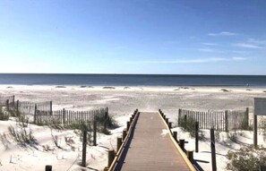 Ein hölzerner Steg führt zu einem Sandstrand unter blauem Himmel.