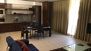 Ein modernes Wohnzimmer mit Sofa, Couchtisch und Fernseher. Es gibt eine Essecke mit Tisch und Stühlen. Der Raum verfügt über eine Küchenzeile mit Mikrowelle und Kaffeemaschine.