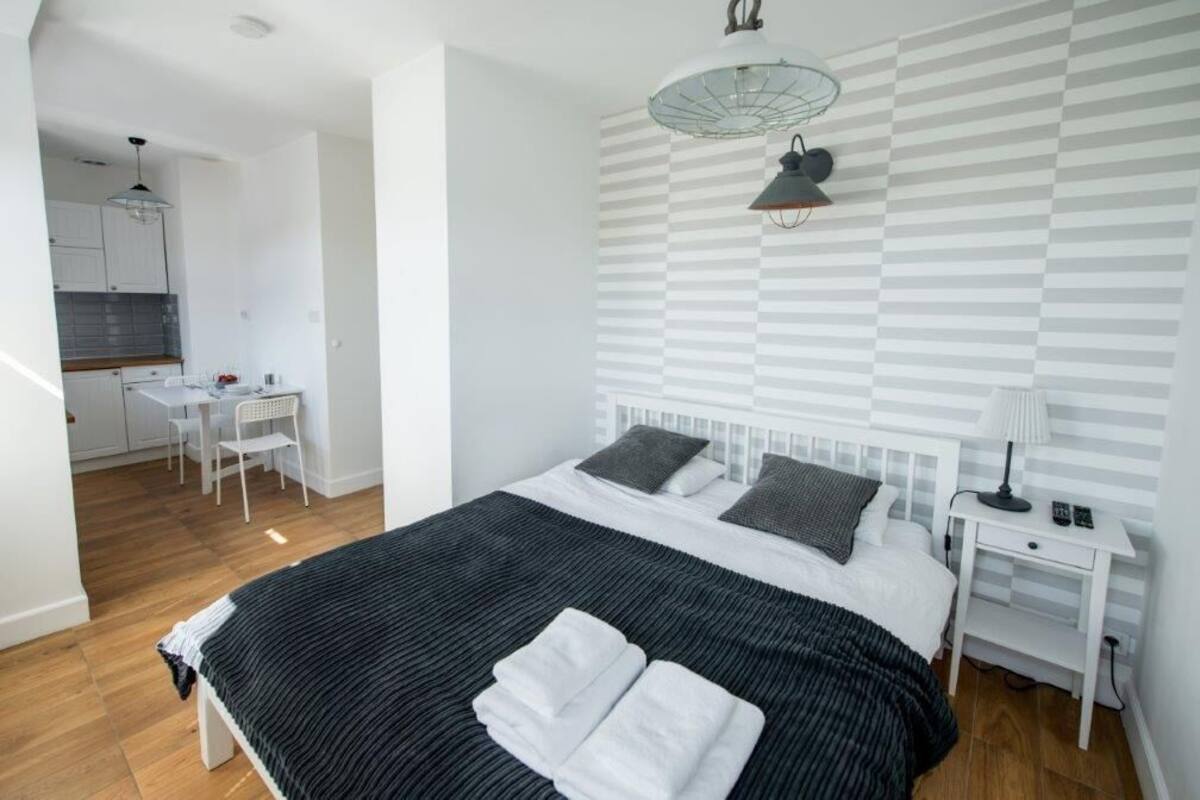 Ein modernes Schlafzimmer mit einem großen Bett, einem Nachttisch, einer Lampe und Blick auf eine Küchenzeile.