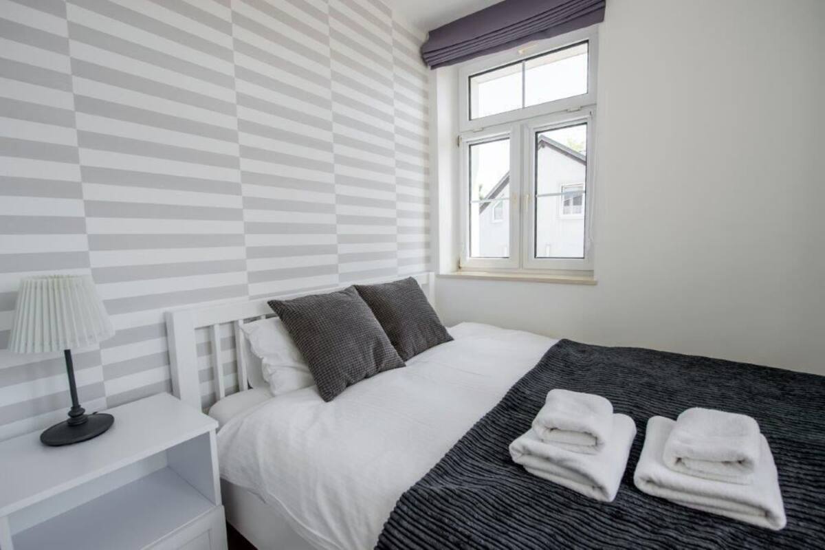 Ein ordentlich eingerichtetes Schlafzimmer mit gestreifter Wand, einem Fenster mit Ausblick, einem Bett mit Kissen, einem Nachttisch mit Lampe und gefalteten Handtüchern auf dem Bett.