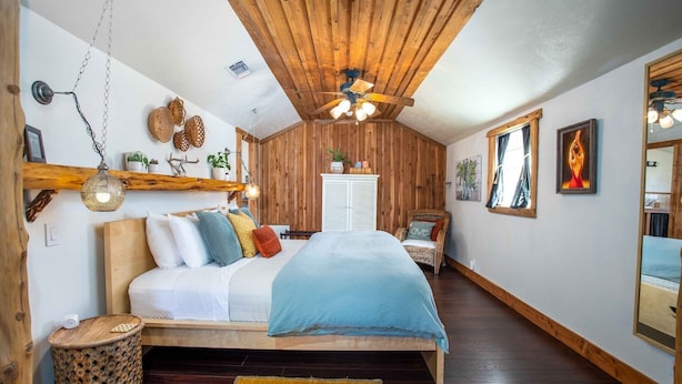 Ein gemütliches Schlafzimmer mit Holzbalkendecke, einem Bett mit vielen Kissen, einem Korbstuhl und einem kleinen Tisch.