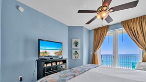 A bedroom with a bed, a television on a stand, a ceiling fan, and a view of the ocean through a balcony. - Gulf-front corner unit. 5th floor. Beach service during season! (Fort Walton Beach)