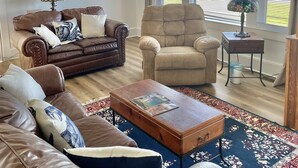 A living room with a brown leather sofa, a beige armchair, and a wooden coffee table. There is a ceiling fan, a window with blinds, and a small side table with a lamp.