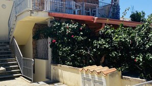 En bostadsbyggnad med en trappa som leder upp till en balkong, en liten trädgårdsyta med blommande växter och en grind av metall.