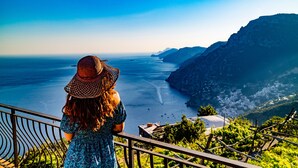 A woman in a patterned dress and wide-brimmed hat overlooking a coastal view with a mountainous backdrop. - Goat Path (Positano)