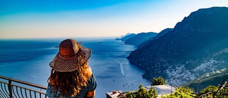 A woman in a patterned dress and wide-brimmed hat overlooking a coastal view with a mountainous backdrop.