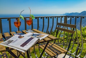 Una mesa de madera con dos vasos de líquido ámbar, un menú y un paraguas verde en un balcón con vistas a un paisaje montañoso.