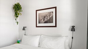 Ein ordentlich gemachtes Bett mit weißer Bettwäsche, ein gerahmtes Schwarz-Weiß-Foto an der Wand, eine hängende Pflanze und zwei Nachttischlampen.