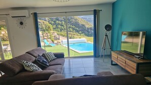 A modern living room with a sofa, a TV, and a view of a swimming pool and mountains.