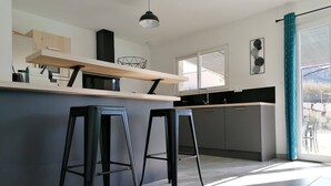 A modern kitchen with a central island, two black bar stools, a hanging pendant light, and a view of the outdoors through a large window.