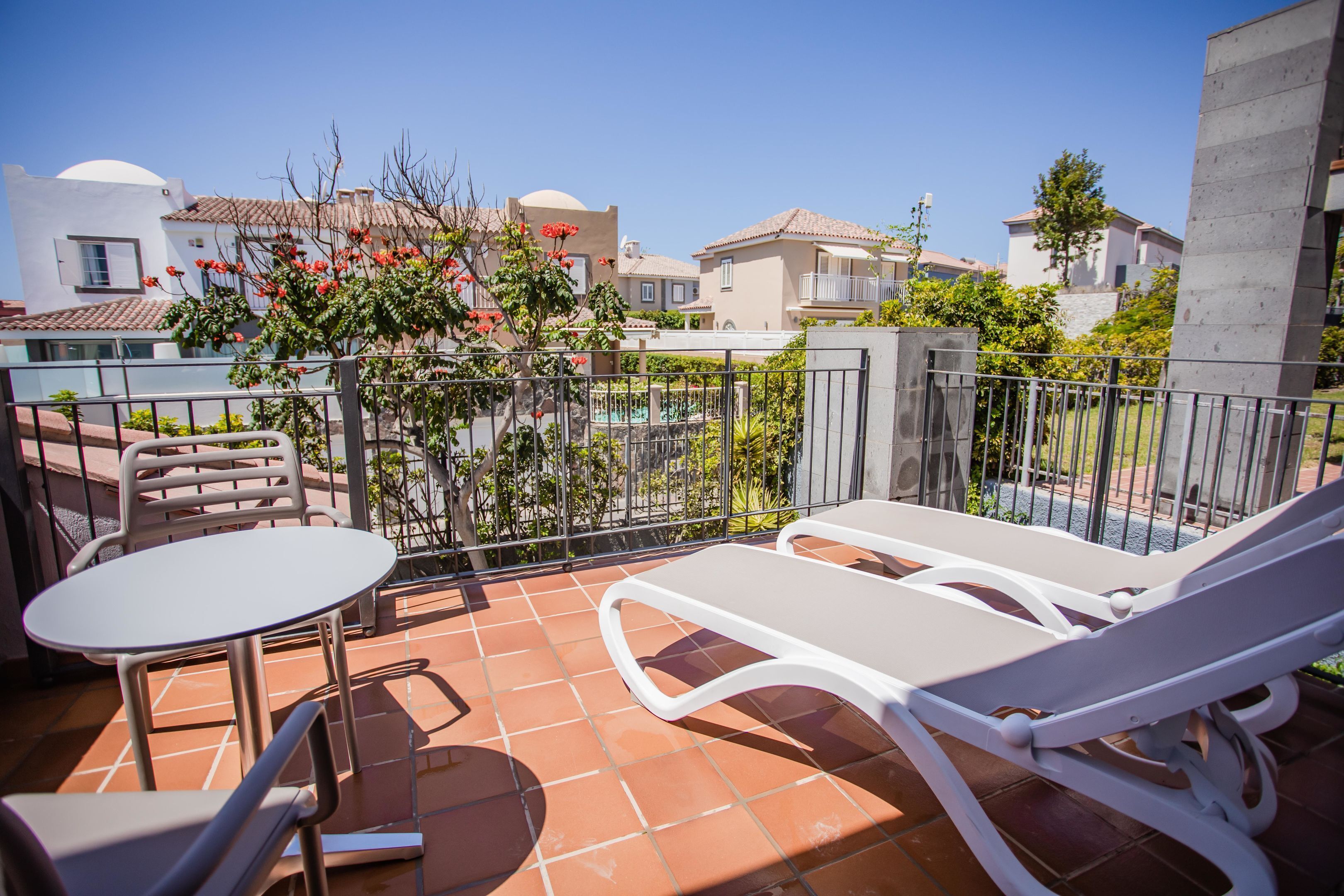 Terraço com mesa, cadeiras e uma espreguiçadeira, com vista para edifícios e áreas verdes.