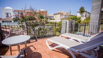 Terraço com mesa, cadeiras e uma espreguiçadeira, com vista para edifícios e áreas verdes.