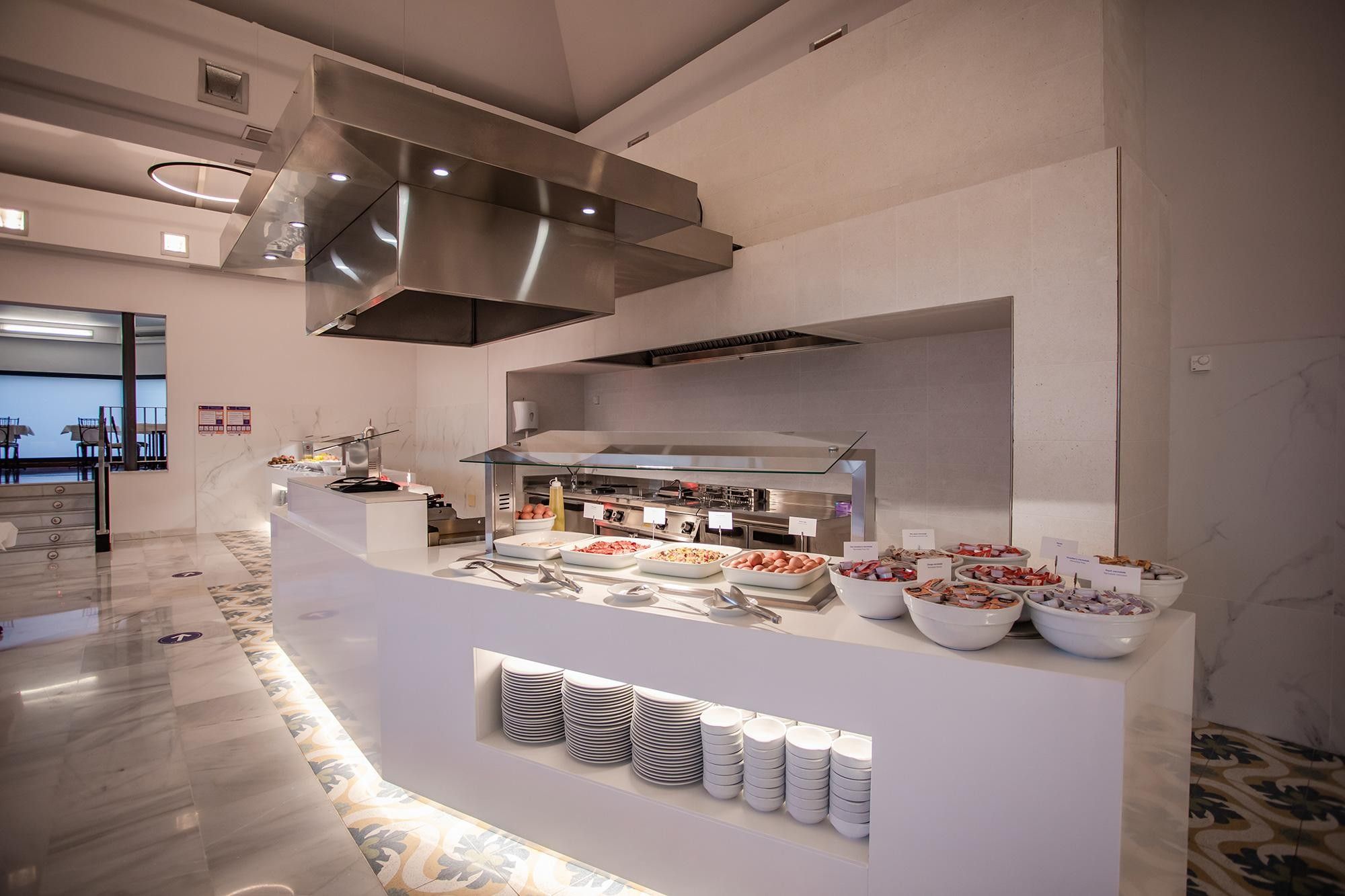 A modern buffet setup with a stainless steel range hood, a glass front display case, and neatly stacked plates and bowls.