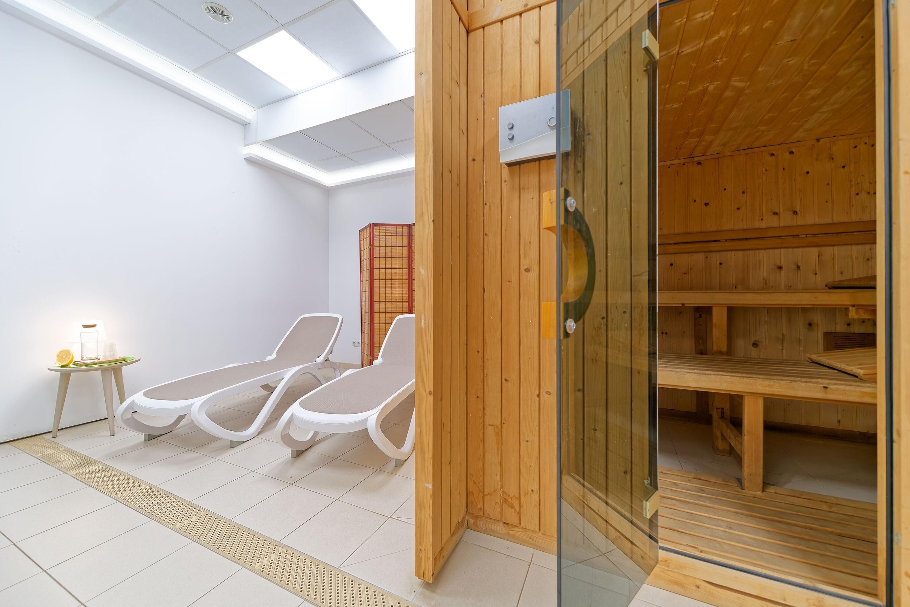 A sauna room with wooden benches and a glass door, a white wall, and a small table with a lamp.