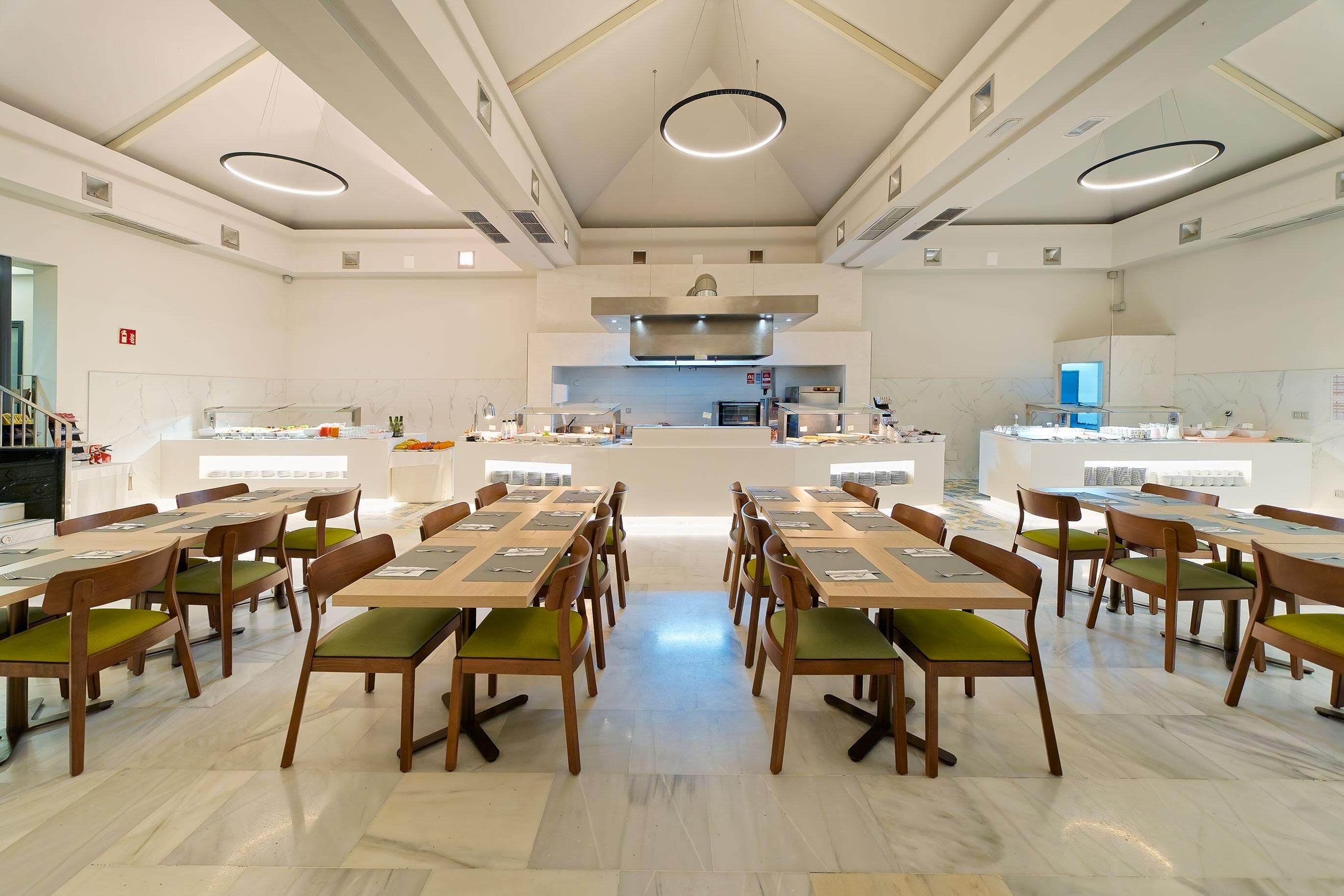 A spacious dining area with multiple tables and chairs, a central kitchen area, and a modern ceiling design.