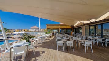A sunlit outdoor dining area with tables and chairs, a pool, and a view of buildings in the background.